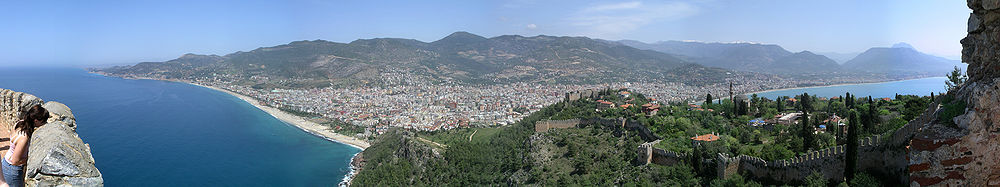 A panoramic view of a city beneath a mountain range with blue sea on both sides of a peninsula. On the peninsula is a castle wall and red roofed buildings. A young girl peers into the scene over the wall on the far left.