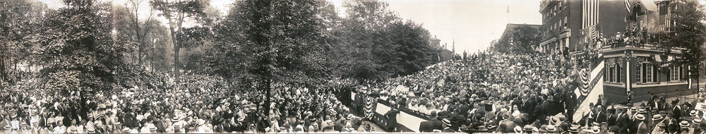 A panorama image showing a large crowd seated in chairs and standing outdoors around a speaker who delivers a speech on a podium in the center. Downtown brick buildings and large campaign posters are in the background.