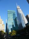 Ground-level view of two tall skyscrapers; the one on the left is shorter and has a blue-green, all-glass facade. The taller building to the right is still under construction; it has an all-glass exterior and a large white spire rising above its slightly tapered roof.