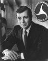 Black and white photo of a man wearing a suit sitting at a desk with his hands folded on it and the DOT logo and US flag behind him