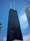 Ground-level view of a 100-story skyscraper with a black exterior and visible X-bracing; the building tapers slightly and is topped by two large antenna masts.