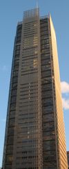 Ground-level view of a 50-story building with a dark glass exterior and a square cross section; a thin spire rises above its roofline.