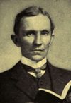  Man, probably mid-forties, dark hair, clean shaven, wearing a high collar with tie, looking staright ahead. He is holding an open book