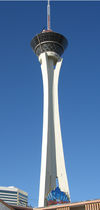 Ground-level view of a tall, concrete tower. The tower curves inward about 30 stories up, but then projects outward. Its uppermost section consists of a rounded, black glass platform and a large, thin spire.