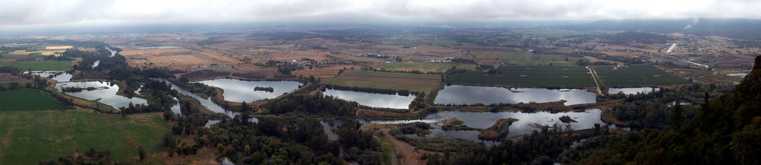 A river, seen from above, winds through flat farmland and a series of ponds on both sides of the stream. Buildings are scattered here and there across the landscape under a cloudy sky.