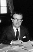 A black-and-white of a man wearing glasses and a suit. He sits inside a house with a hand placed on a table with a pile of papers.