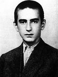 A black-and-white photograph of a young man looking at the camera. He has short, dark hair, thick dark eyebrows, and is wearing a dark, buttoned-up jacket and light shirt.