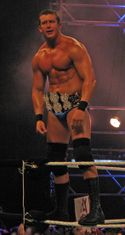 A dark-haired man wearing blue wrestling tights, black knee-high wrestling boots and black knee-pads stands on the white ropes of a wrestling ring. He is wearing around his waist a gold-plated professional wrestling championship belt shaped with three dollar signs on the front.