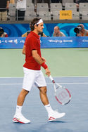 A dark-haired man is in a red shirt with white shorts and shoes and bandanna, which he is carring his tennis racket in his right hand pointing towards the ground