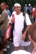 A dark-haired man in all white clothing, and caring a redish-black bag on his right shoulder and a black one on the left shoulder