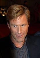 A blond-haired man with blue eyes wears a black jacket with a grey shirt.