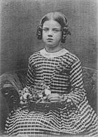 Three quarter length studio photo of seated girl about nine years old, looking slightly plump and rather solemn, in a striped dress, holding a basket of flowers on her lap.