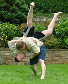 Two men in combat appear on a green lawn in front of a  shrubbery. Both men are wearing dark coloured shorts and cream coloured tops. One man has a tactical advantage, and is throwing his opponent head-first to the ground.