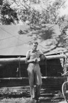 Whitlam in military uniform stands under a tree in front of a large tent.  He holds a mug in his hand.