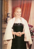 A picture of a Evita who is blond lady and former strongman/woman of Argentina. Her hair is drawn into a tight bun at the back. She is wearing a black, low-cut dress. Around her neck is a number of chains. The lady's hands are folded in her front and she has a white fur shawl around her. Behind her, a red curtain and flowers are visible