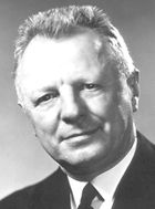 Head and shoulder of a 60-ish man with a flattop haircut and in a coat and tie, looking directly at camera with head tilted to his right and a slight smile.