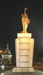 A stone statue of torch-bearers as seen at night. A fountain with a white base is in the background