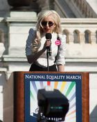 A blond woman speaking on a kiosk. She wears a white shirt and black glasses. Behind her, the balcony of a building is visible.