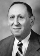 Head and shoulders of a man in his early 60s in coat and tie, facing slightly to his right. He is balding and has a serious but slightly smiling expression.