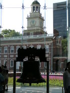 The Liberty Bell hangs in a glass-backed structure, with a brick, 18th century building with a steeple visible in the background.