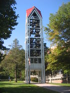 A three-dimensional vertical structure perhaps 70�feet (21 m) tall and 10�feet (3.0 m) wide by 10�feet (3.0 m) long is filled with tiers of large metal bells. The structure, standing in front of a large institutional building in a park-like setting, is supported by several metal legs. A sidewalk passes through the structure, which is open at the bottom to a height of perhaps 10�feet (3.0 m).