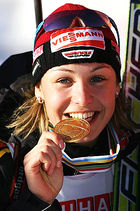 A woman in predominately black winter sportswear and a red cap smiles into the camera while biting into a gold medal.