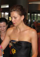 A brown haired woman looking away from the camera. Her hair is tied back, and she is wearing gold earrings and a shoulderless, sleeveless black dress with a yellow, red and blue patten on it.