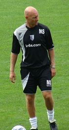 A bald man in a black T-shirt and shorts with grey and white trim, with a football at his feet.