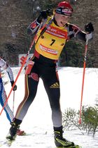 A woman cross country skis uphill towards the camera. She wears black winter sportswear and a bright red jersey with the number 13. A second skier behind her can be seen on the right.