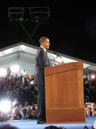 Obama delivers a speech at a podium while several flashbulbs light the background.