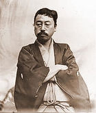 Portrait of an Asian man with  moustache dressed in traditional Japanese cloths. He is looking down  with his arms crossed.