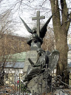 A statue of a winged angel holding a large stone cross over the bust of a balding middle-aged man.