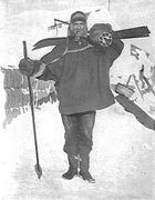  Man, standing, wearing a smock, heavy trousers and boots. He has a ski stick in his right hand, a pair of skis strapped on his back, and is carrying a rounded bundle on his shoulder. Behind him on the ground is assorted polar equipment.