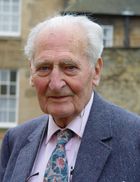 A white elderly man wearing suit and tie stands before a brick house