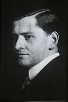 A black and white portrait of a man in a suit and tie. Half of his face is in a shadow.