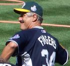 An older white male wearing a green cap with a yellow bill, a blue jersey, with the lettering "FINGERS" and a number 34 below it on the back, on a grass field.