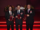 Three white men wearing suits and ties stand in front of a podium, each holding up a medal