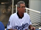 A gray-haired white male, wearing a white uniform with "Dodgers" across it, sitting in a bullpen with a white fence in the background.