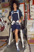 A woman wearing a blue t-shirt and shorts, seen running on a treadmill to which she is attached by white strapping. Floating equipment and cabinets are visible in the background.