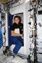 An astronaut emerges from a small sleeping compartment surrounded by equipment.