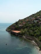 A hill populated with houses slants down into a blue-green sea where a single boat floats in front of an stone dock with five arches. A stone wall extends along the sea from the dock to the lower right.