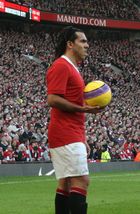 A man wearing a red football shirt, white shorts and black socks. His hair is held back by a hairband. He is holding a yellow football with purple detailing.