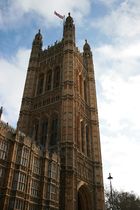 View from below of a large square tower, connected at the left to a lower building. It features an arched portal at its base, and two rows of three high windows on each side. The corners are formed by round turrets, which rise above the main roof-line. A flag flies from a pole at the top of the tower.