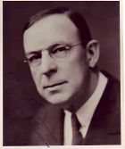 Upper body of a well-dressed man, who is wearing glasses and a coat and tie, with a large forehead and dark hair combed to the side