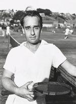 A black-haired man in a white polo shirt poses for a portrait, looking into the camera as he stands next to a tennis net, holding a wooden racket