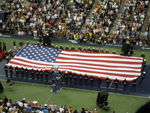 Around thirty soldiers hold a giant American flag on a blue and green hard court, surrounded by crowded bleachers