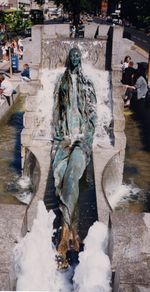 Figure of a young woman sitting on a slope with legs crossed. It is in the middle of a rectangular fountain, surrounded by flowing water.