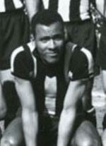 A young man sitting down, wearing a striped shirt. Behind him, three men wearing the same shirt and dark shorts are partially visible