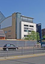 A quirky modern five-storey building with a large sign saying "BBC Yorkshire" in black above the second-floor windows on the white-fronted facade of the lower four floors can be seen on the far side of a dual-carriageway road with a barrier along the central reservation.  At right-angles to the right of the building is a tall blue slab with the letters "BBC" in white at the top.  The left side of the building is mostly brick-red with a few windows, but above it is a light blue windowless section.  The roof above this and the grey fifth floor of the frontage curves gently down to the rear.  A lone car is driving from left to right along the road;  between it and the building, temporary boards have been erected in front of a building to the left.  In the top left-hand corner of the picture, part of a tall many-windowed building can be seen.