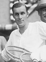 A black-haired man in a white shirt, holding several wooden rackets under his arm